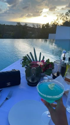 a person holding up a drink at a table by the water with plates and utensils on it