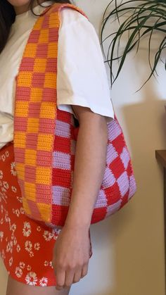 a woman is holding a crocheted bag in her right hand while standing next to a potted plant