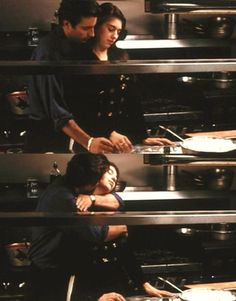 two people in a kitchen preparing food on pans and stove top burners, with one person leaning over the counter