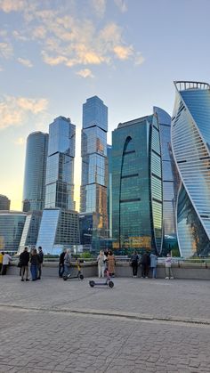 many people are standing around in front of some very tall buildings and skateboarders