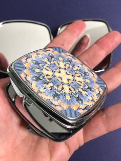 a hand holding a compact case with a flower design on it and two mirrors in the background