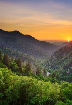 the sun is setting in the mountains with trees on each side and hills to the left