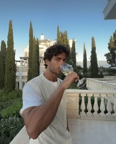 a man drinking from a wine glass on top of a balcony with trees in the background