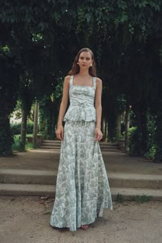 a woman standing in front of some trees wearing a green and white dress with tiered ruffles