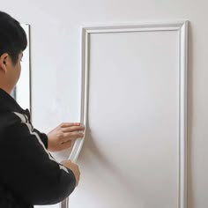 a man in black jacket painting a white frame on wall with paint rollers and brush