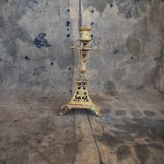 an ornate gold candle holder stands in front of a gray wall with peeling paint on it