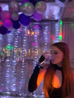 a woman with long red hair drinking from a wine glass while standing in front of balloons