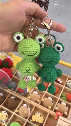 two crocheted frog keychains are being held in front of other stuffed animals