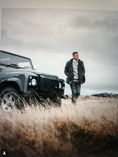 a man standing next to a black land rover in a field with tall brown grass