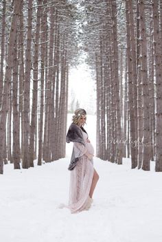 a pregnant woman is standing in the snow by some trees and wearing a fur coat