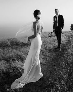 a bride and groom walking in the grass on top of a hill with their veil blowing in the wind