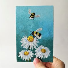 a hand holding up a piece of paper with bees and daisies on it in front of a blue background