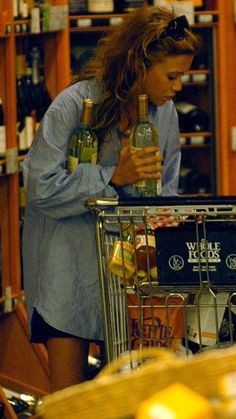 a woman holding a bottle in her hand while standing next to a shopping cart filled with wine bottles