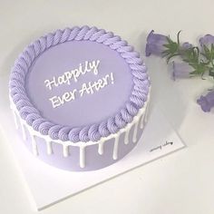 a purple birthday cake with white icing and flowers on the table next to it