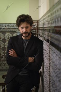 a man leaning against a wall with his arms crossed and looking at the camera while wearing a suit