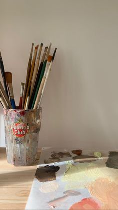 a cup filled with lots of paint brushes on top of a wooden table next to an easel