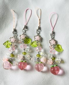 four charms are hanging on a white sheet with pink and green beads, one is shaped like an apple