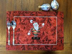 a red place mat with a santa clause design and two silver spoons next to it