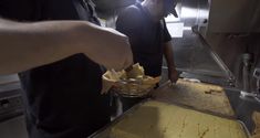 two men preparing food in a kitchen with metal trays on the counter and one man holding a basket