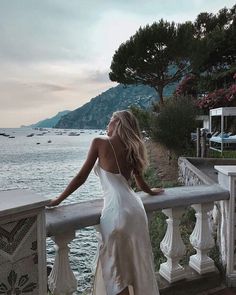 a woman in a white dress is standing on a balcony overlooking the water and mountains