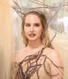 a woman with long blonde hair wearing a dress and holding branches on her head, standing in front of sheer curtains