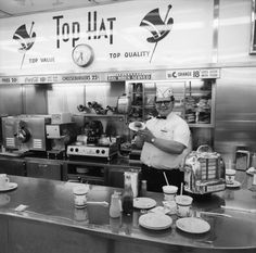 a man standing in front of a counter with plates and cups on it, next to a sign that says top hat