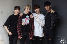 four young men standing next to each other in front of a gray wall with one pointing at the camera