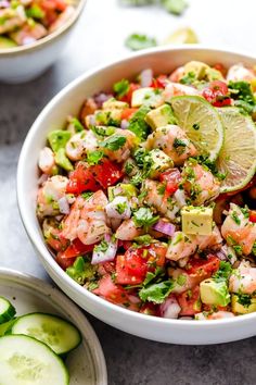 a white bowl filled with shrimp salad next to cucumbers and lime wedges