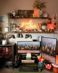 Cozy desk setup with dual monitors. A fall forest as the screen background. Two shelves above the desk with a neon sign that says Good Vibes and cozy gaming decor. Cluttered Gamer Room, Cozy Office Game Room, Gaming Setup Organization, Cozy Pc Gaming Setup, Eclectic Gaming Room, Cozy Gamer Office, Cosy Games Room, Maximalist Gaming Room, Boho Game Room Ideas