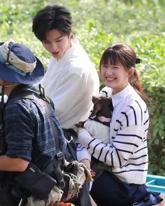 three people sitting on the ground with one holding a small dog and smiling at the camera