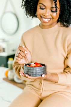 a woman sitting on a bed holding a bowl of strawberries in her hand and smiling at the camera