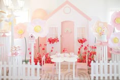 a doll house with pink and yellow flowers in the front yard, surrounded by white picket fence