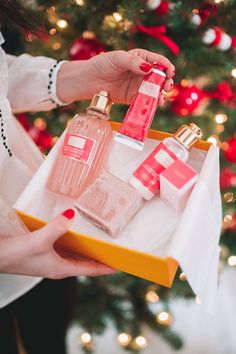 a woman holding a box with three different bottles in front of a christmas tree