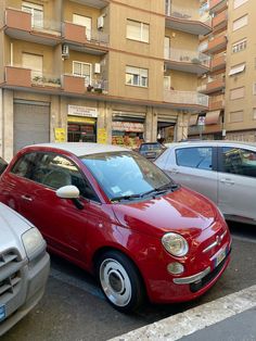 a small red car parked next to other cars in a parking lot near tall buildings