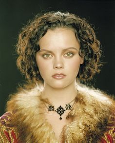 a woman with curly hair wearing a fur collar and necklace on her neck is looking directly into the camera