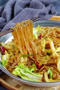 a bowl filled with noodles and vegetables being held by chopsticks over the top