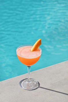 an orange drink sitting on top of a table next to a swimming pool with water in the background