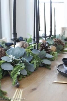 a wooden table topped with candles and greenery next to a candle holder filled with pine cones