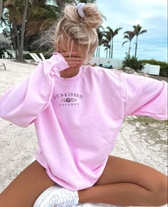 a woman sitting on the beach wearing a pink sweatshirt with blue writing that says god is for focus, need a palm tree