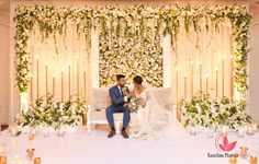 a bride and groom sitting on a couch in front of a floral wall with candles