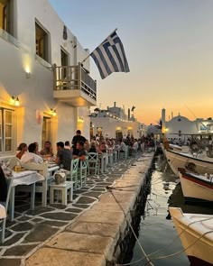 people are sitting at tables near the water in front of boats and buildings with lights on them