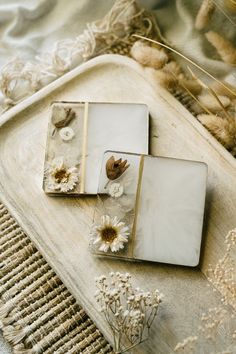 three square glass coasters with white flowers on them sitting on a table next to dried grass