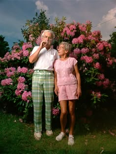 an older man and woman standing next to each other in front of pink flowering bushes