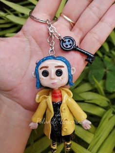 a hand holding a key chain with a small doll on it's thumb and wearing a yellow coat