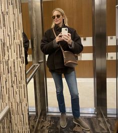 a woman standing in an elevator taking a selfie with her cell phone while wearing jeans and boots