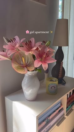 a white vase with pink flowers sitting on a table next to a lamp and books