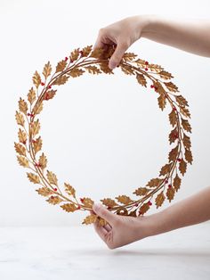 a person holding a wreath made out of leaves and berries on a white table top