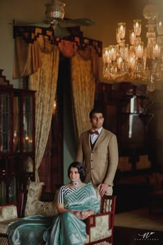 a man standing next to a woman sitting on a chair in front of a chandelier