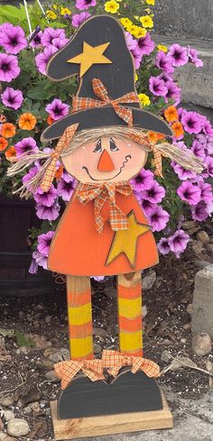 a wooden scarecrow standing in front of purple and yellow flowers with a star on its head