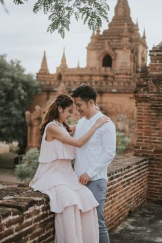 a man and woman standing next to each other in front of a brick wall with an old building behind them